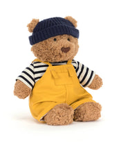 Teddy bear wearing a navy hat, striped shirt, and yellow overalls on a white background