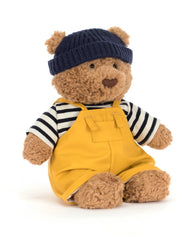 Teddy bear wearing a navy hat, striped shirt, and yellow overalls on a white background