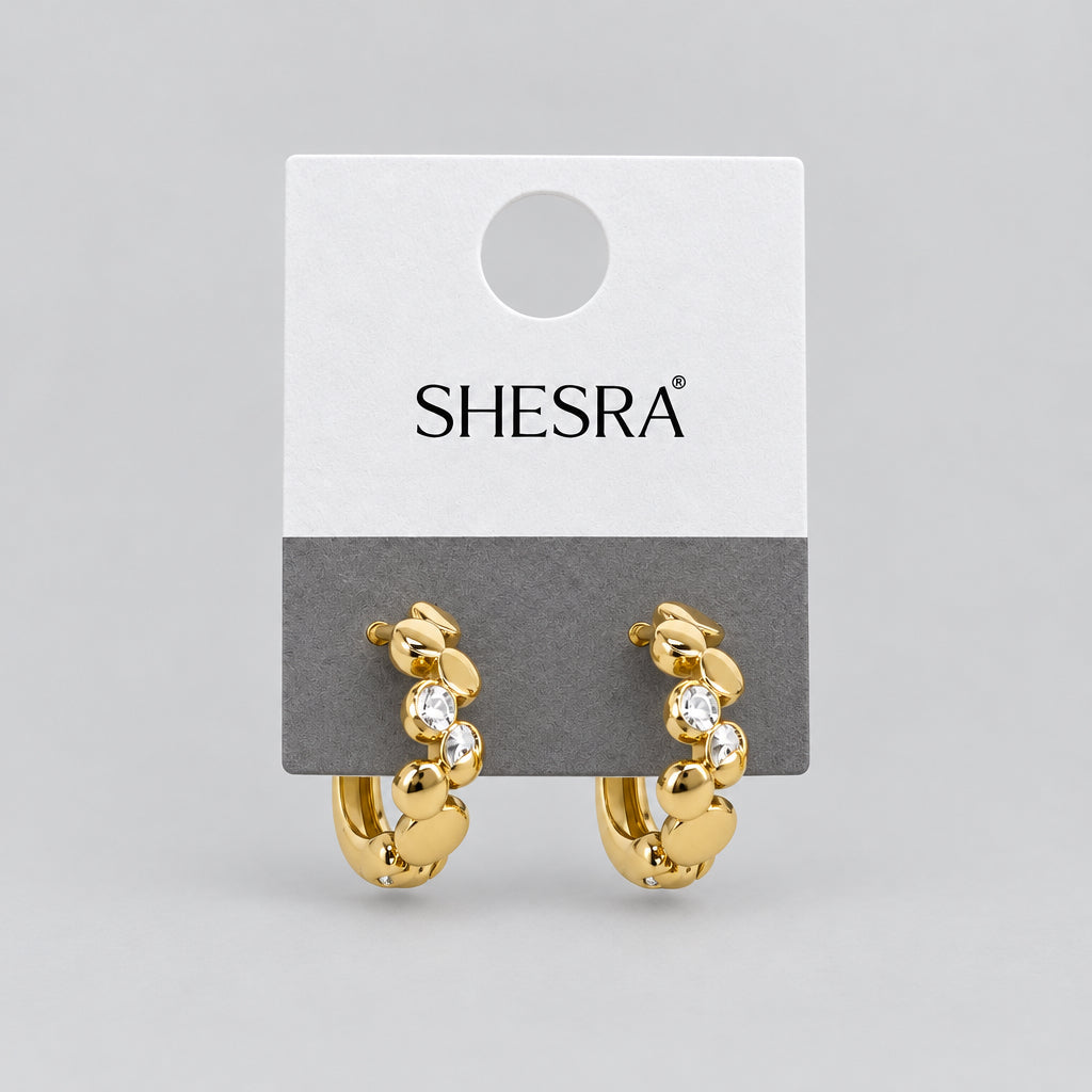 A pair of gold crescent-shaped hoop earrings with small clear rhinestone accents, attached to a white and gray display card printed with the brand name 'SHE'S RA'.