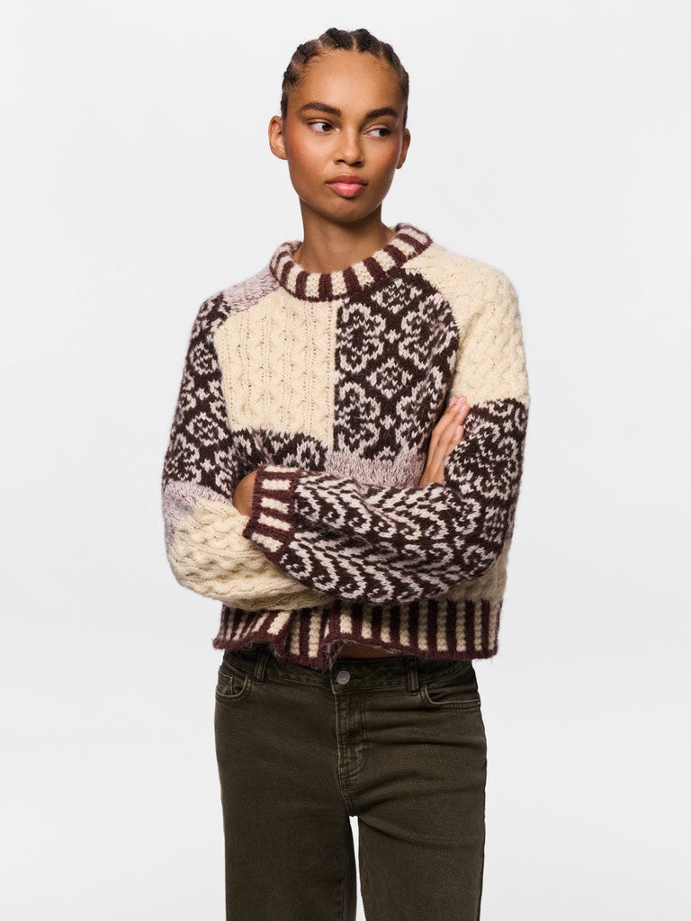 A woman modeling a chunky knit crewneck sweater with cream and dark brown Fair Isle patterns, paired with dark olive green jeans, against a plain light gray background.