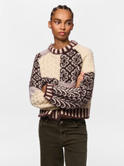 A woman modeling a chunky knit crewneck sweater with cream and dark brown Fair Isle patterns, paired with dark olive green jeans, against a plain light gray background.